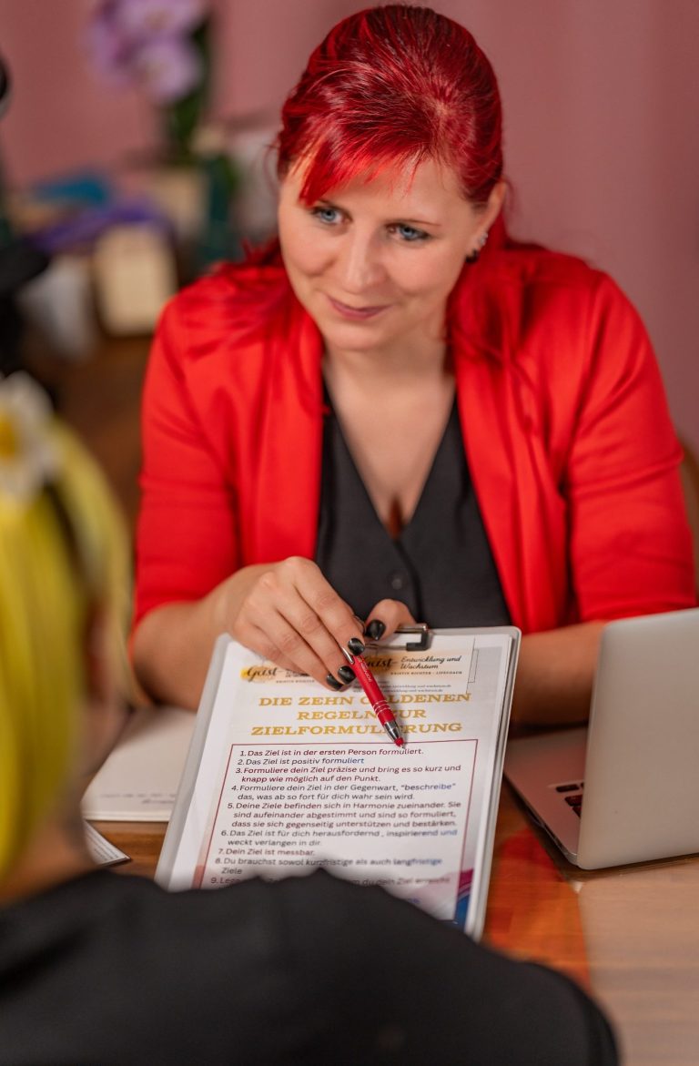 Eine Person mit rotem Blazer hält ein Notizbuch und einen Stift in der Hand.
