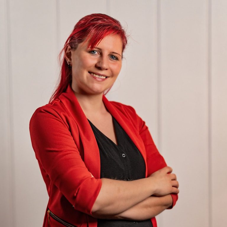 Frau mit rotem Blazer und roten Haaren, lächelnd mit verschränkten Armen.