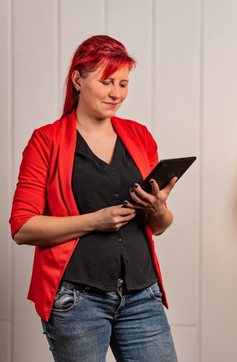 Frau mit roten Haaren und rotem Blazer, die ein Tablet in der Hand hält.