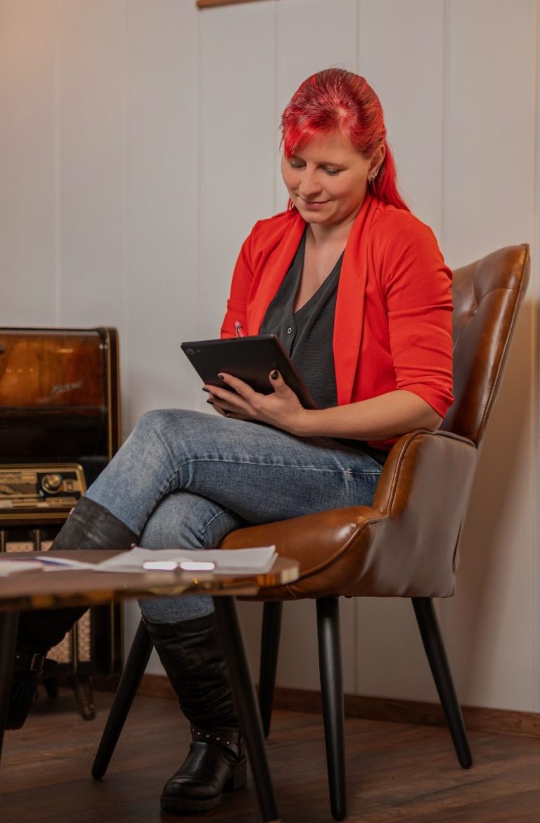Frau mit roten Haaren und rotem Blazer, die ein Tablet in der Hand hält.